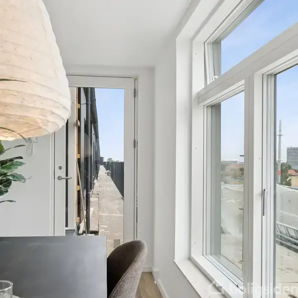 Et lyst rum med en lampe hængende over et bord, hvor der står glas. Rummet har store vinduer med udsigt til en altan og bylandskab udenfor.