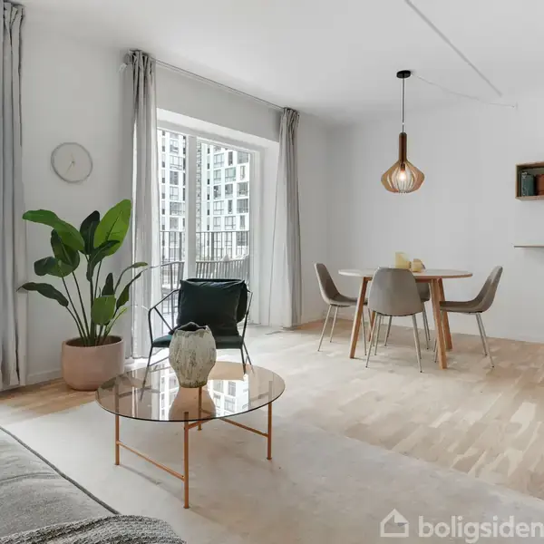 Stue med runde bord og vase i forgrund, stående på lyst trægulv. I baggrunden ses spisebord med stole, hængende lampe og hylde. Store vinduer viser bybygninger udenfor.