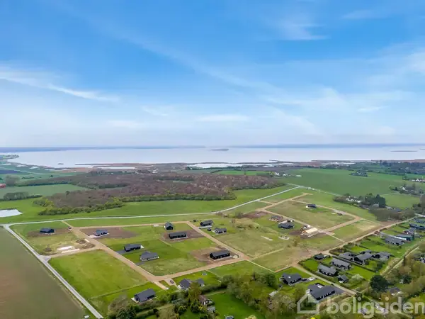 Landskabet viser et åbent område med flere spredte huse under opførelse, omgivet af marker og skov, med udsigt over en fjord eller sø i baggrunden under en klar blå himmel.