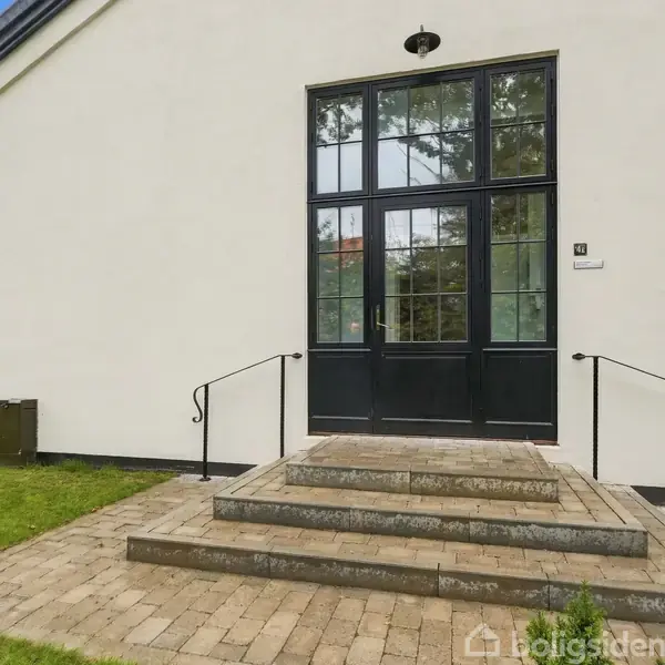 Dobbeltdør af glas åbner mod en stentrappelanding med tre trin, flankeres af sorte gelændere. Græsplæne og hvid bygning med tegltag i baggrunden.