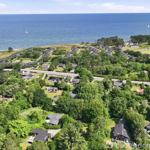Kystnært boligområde med grønne træer og huse spredt ud i landskabet. Et stort hav strækker sig i baggrunden, mens enkelte sejlbåde ses på vandet.