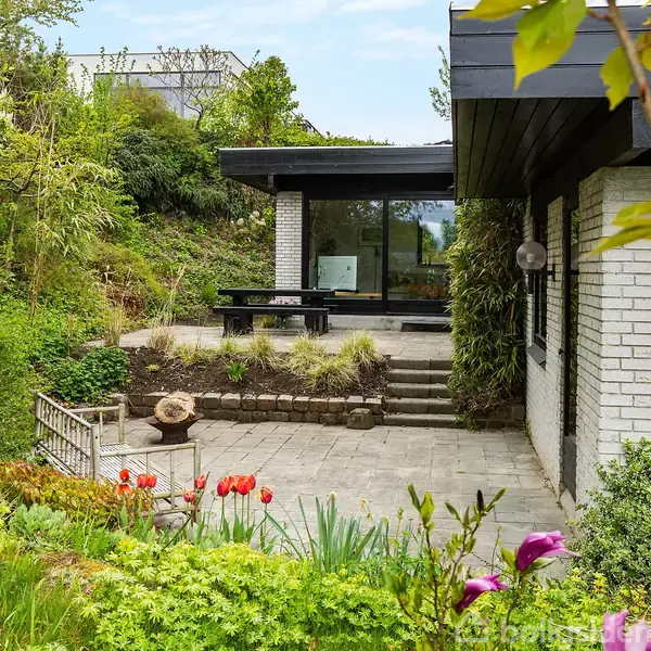 Et hus med hvid murstensfacade og sorte detaljer. Foran huset er der en flisebelagt terrasse med en bænk og blomsterbede. Omgivende have med grønne buske og blomster.