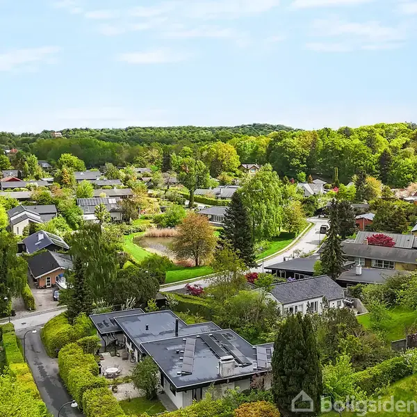 Beboelsesområde med huse spredt ud i frodige grønne omgivelser. Træer og buske omringer bygningerne, mens en snoet vej forbinder dem. Rolig atmosfære og naturskønne omgivelser.
