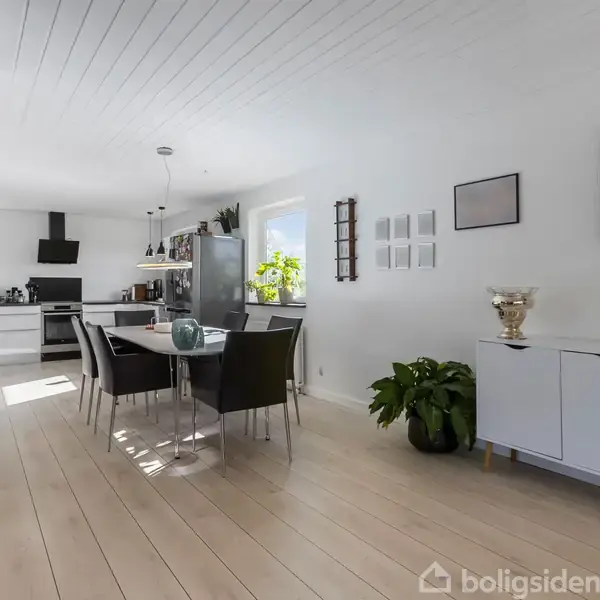 Køkken-alrum med spisebord og sorte stole i midten. Hvidt interiør og træloft skaber en lys stemning. Planter og kunst på væggen, skaber en hyggelig atmosfære.