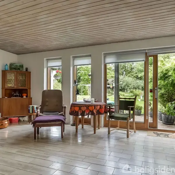 Stue med brændeovn til venstre. I midten er der en lænestol og et rundt bord med dug. Store vinduer og havedøre åbner ud til grøn have.