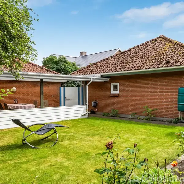 En liggestol står på en velplejet græsplæne. Et bord med stole under en overdækket terrasse. Baggrunden domineres af et rødt murstenshus med brune tagsten.