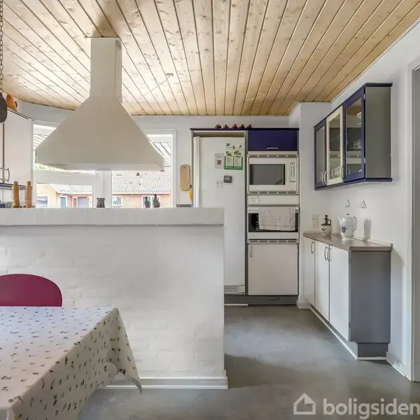 Køkken med hvid emhætte over gråt køkkenbord, omgivet af hvide skabe og apparater. Et bord med dug og røde stole står ved siden af en rød dør. Lofter i lyst træ.
