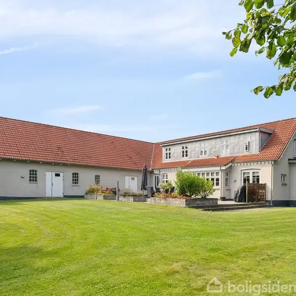 Et hvidt landhus med rødt tegltag står på en velplejet græsplæne. Huset har en terrasse med blomsterbede og er omgivet af træer under en klar blå himmel.