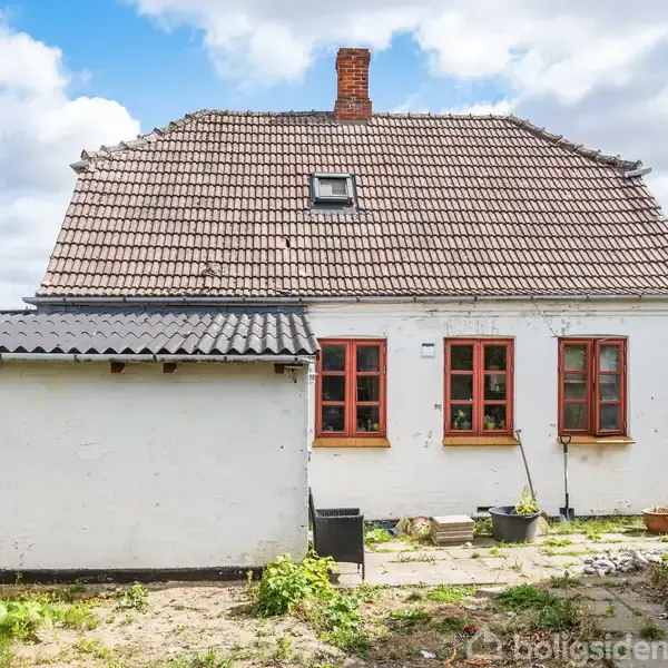 Hus med hvid facade og rødt tag ligger i en have. Murstensskorsten stikker op fra taget. Potteplanter og små buske omgiver huset. Sollys skaber en lys atmosfære.