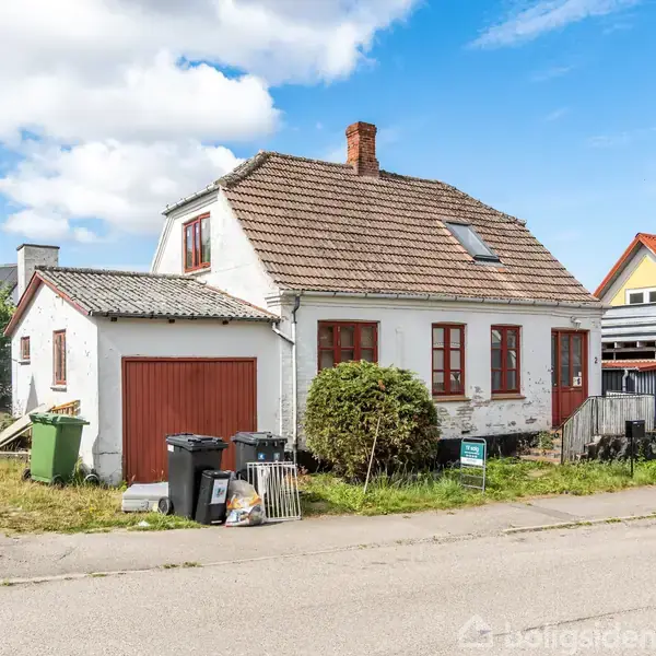 Hus med hvid facade, rødt tegltag og røde vindueskarme, står langs en vej. Til venstre ses affaldscontainere og en garage. Omkringliggende huse har farverige tage.