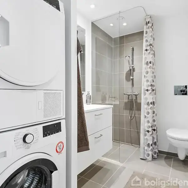 Vaskemaskine og tørretumbler står stablet i et moderne badeværelse med bruser og toilet. Brusenichen har et gråt flisegulv og et bruseforhæng med mønster.