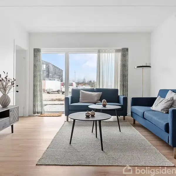 To blå sofaer står i en modern stue. Et lavt bord er placeret på et lyst tæppe på trægulvet. Store vinduer slipper lys ind.