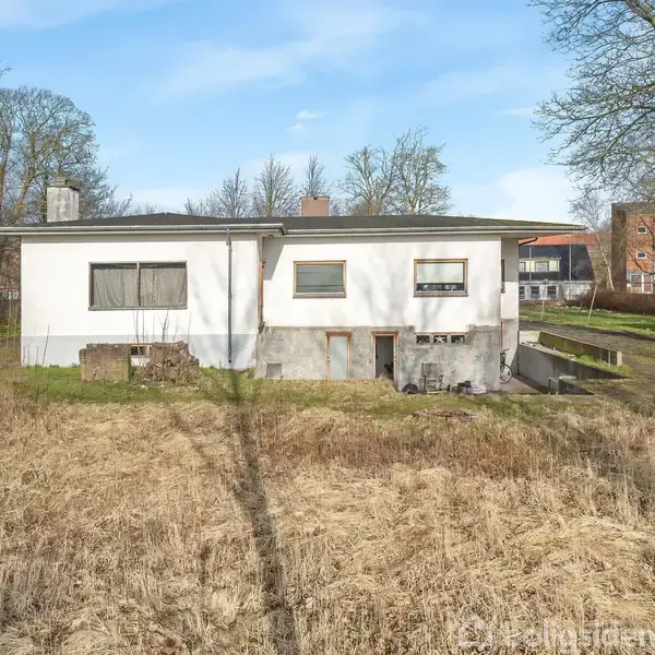 Et hvidt, rektangulært hus med fladt tag står på en åben, tør græsmark. Der er træer i baggrunden og en garage til højre.