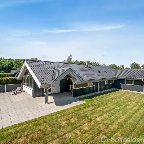 Et enplanshus med grå tagsten og lejlighed, ligger i en velplejet have med grønt græs. Foran huset er der en stor flisebelagt terrasse omgivet af hække.