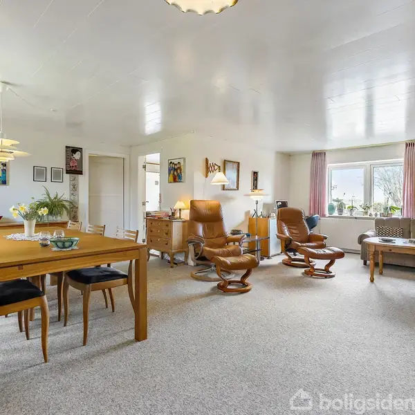 Spisestue med et langt træbord og otte stole, omgivet af lys, rummelig stue med brune lænestole, sofabord, sofa og store vinduer indrammet af lyserøde gardiner.