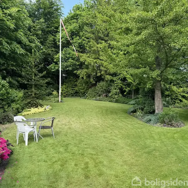 En græsplæne med et lille bord og to stole står på en velplejet græsplæne omgivet af blomsterbede og store træer. En flagstang rejser sig i baggrunden.