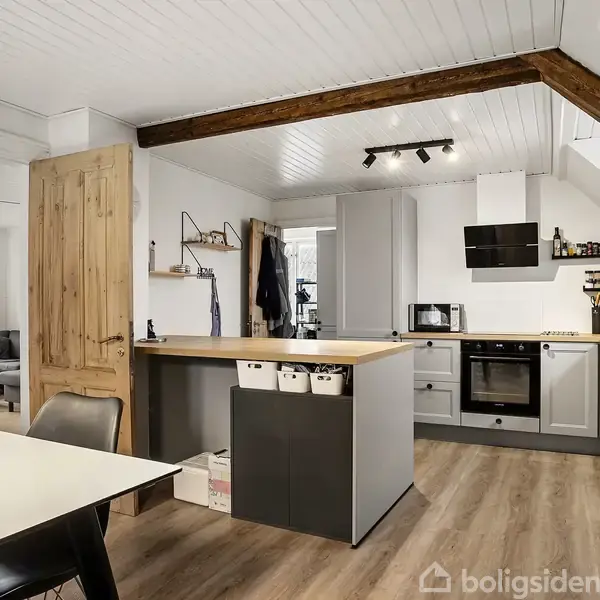 Et moderne køkkenalrum med grå skabe og træbordplader. En åben dør og spisebord forbinder til stueområdet, hvor sofaer ses. Loftet har en synlig bjælke og skråvindue.