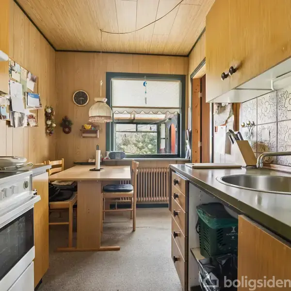 Køkken med ovn til venstre, vask og skab på højre side. I baggrunden et spisebord med stole ved vindue, omgivet af træpaneler og blomstrede fliser.
