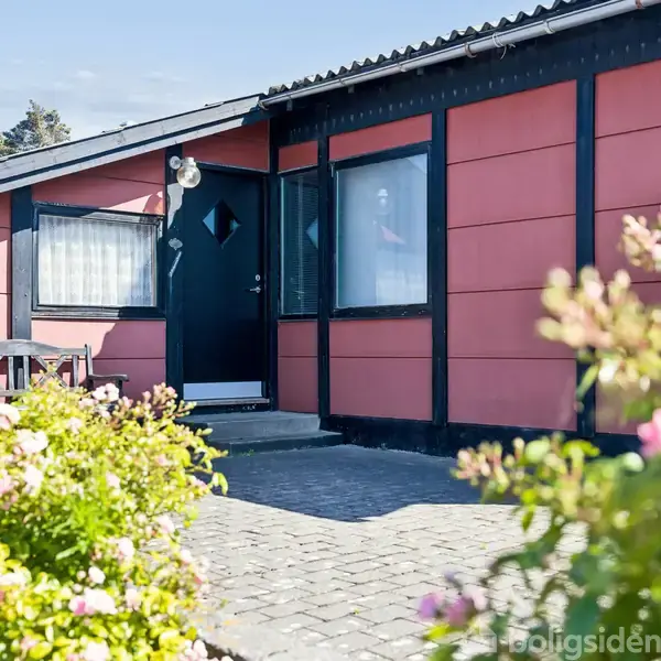 Et rødt hus med sorte detaljer står foran en brolagt indgang. En sort dør er i centrum, omgivet af vinduer. Forgrunden er fyldt med blomstrende buske og en blå himmel ses øverst.