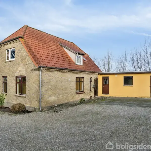 Et gulstenshus med rødt tegltag står på en grusbelagt gårdsplads. Vedhæftet er en lavere bygning i gul med en brun dør. Baggrund med træer og mark.