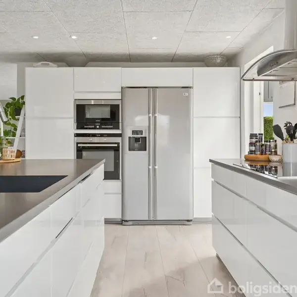 Et moderne køkken viser et stort, dobbeltdørs køleskab integreret i hvide skabe. En køkkenø med vask og planter suppleres af en kogeplade ved siden af køkkenredskaber.