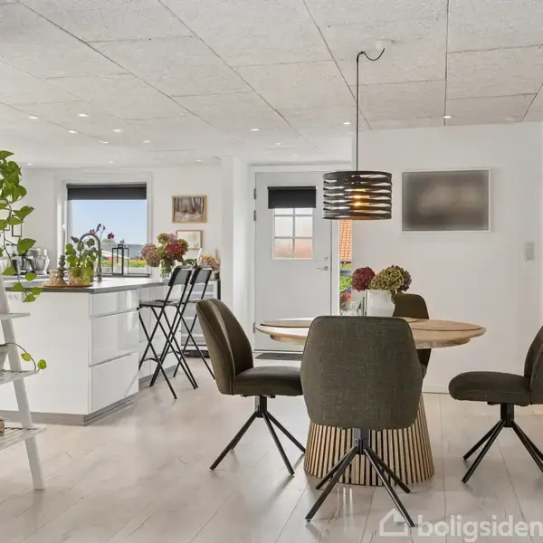 Stue med spisebord og fire stole i moderne indretning. Rundt bord med lampe hængende over sig. Køkkenø med barstole i baggrunden. Grønne planter pynter rummet.