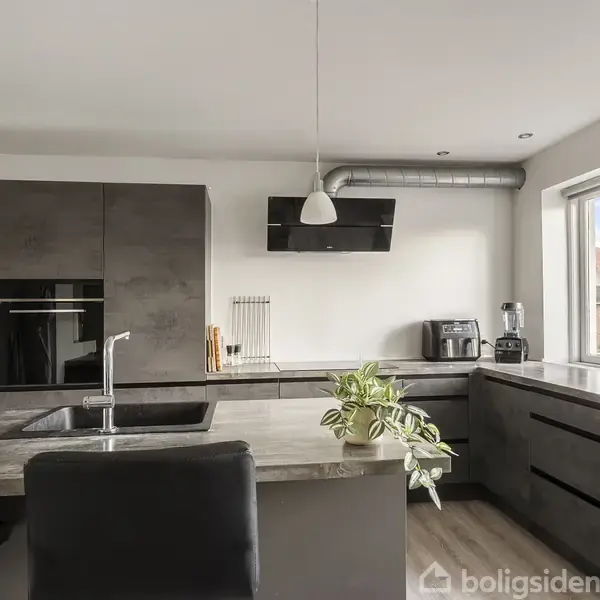 Et moderne køkken med mørkegrå skabe og bordplader. Der er en indbygget ovn, vask med armatur og køkkenudstyr på bordet. Et stort vindue til højre slipper lys ind.