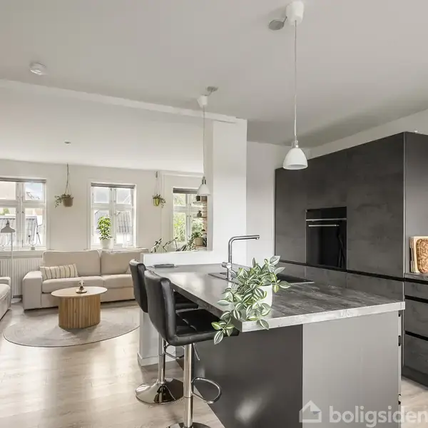 Et moderne køkken med en stor ø af marmor med barstole, indbygget ovn og skab. Ved siden af er en lys stue med sofaer, sofabord og vinduer med planter.
