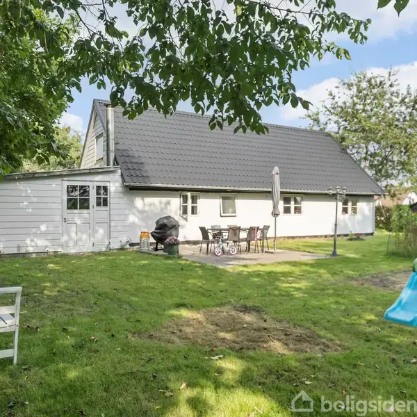 Hus har sort tag og hvide vægge, står i solrig have med rutsjebane, parasol, grill, havebord og -stole. To bænke står på græsplæne under skyggefulde træer.