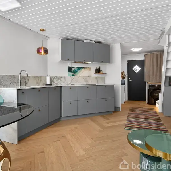 Køkkenet har grå skabe og en marmorarbejdsplade, med hylder over og en malet væg. Rundt spisebord i glas og stole til venstre, en grå sofa til højre.