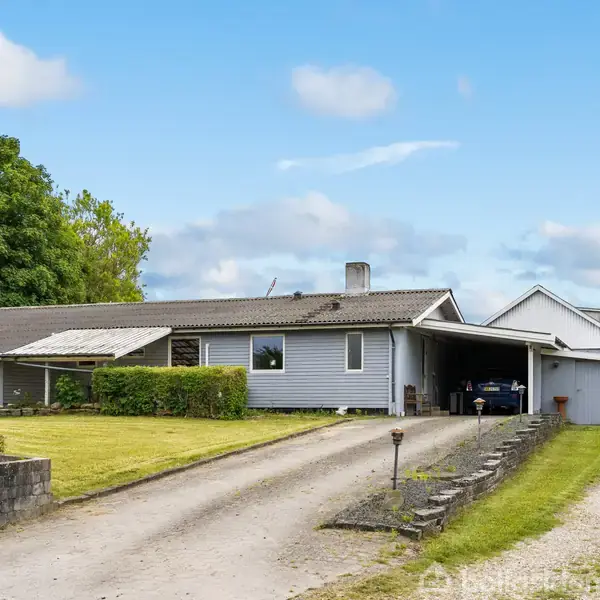 En grå, én-etagers villa med carport står ved en grusvej omgivet af grønne træer og en græsplæne. Skilt foran huset viser teksten "Til Salg".