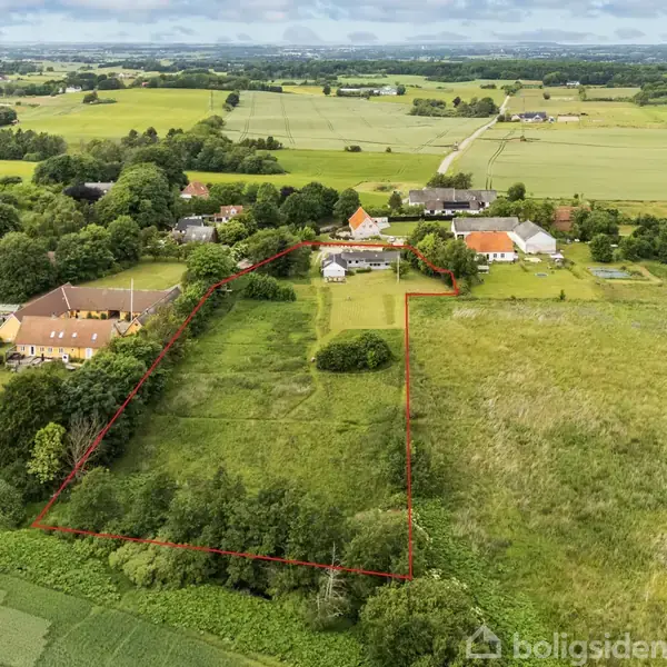 Luftfoto af et stort, grønt landområde indhegnet med røde linjer. Flere huse ligger spredt i landskabet omgivet af træer og marker under en overskyet himmel.