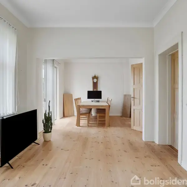 Et minimalistisk rum med et trægulv. En fladskærm og en kaktus står ved vinduet. I baggrunden er et spisebord med stole samt en vægur.