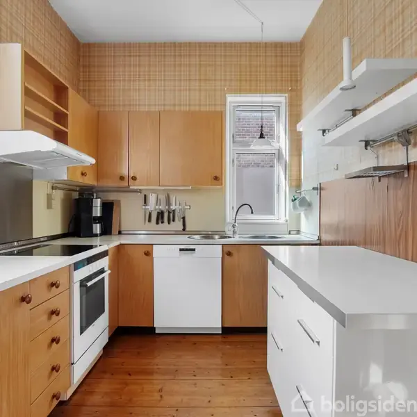 Køkken med træskabe og -skuffer, hvide bordplader, og hvidevarer som ovn og opvaskemaskine. Hylder og kroge på væggen. Vindue med gardin giver lysindfald. Ingen tekst fundet.