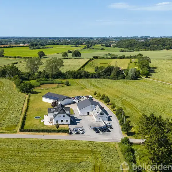 Hus ligger stille ved landevej omgivet af frodige marker og en grøn skov i det fjerne. Flere biler er parkeret nær bygningerne.