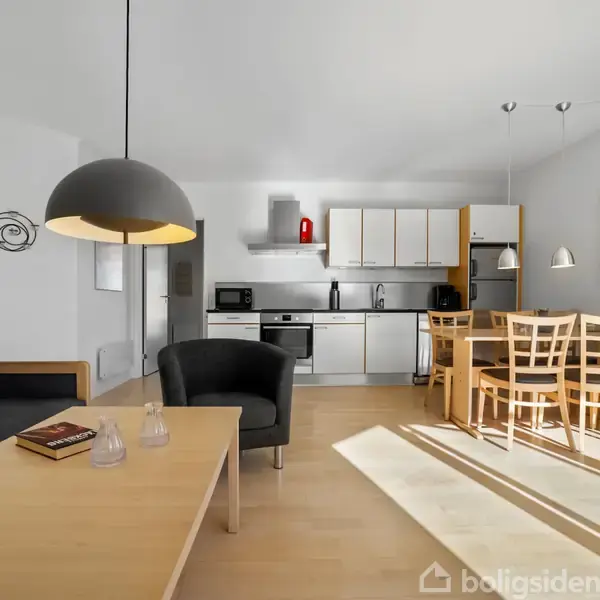 En stue med åben køkken, som indeholder et spisebord med seks træstole. Sofa og sofabord står ved siden af en sort lampe. Kunst er hængt på væggen.
