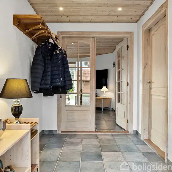 Gang med jakker hængende på en knagerække, en bordlampe på et lille bord, og en glasdør åbner til stuen. Flisegulv og træloft skaber en hyggelig atmosfære.