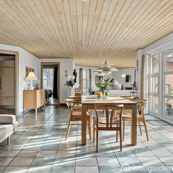 Et spisebord står i midten af et lyst, rummeligt rum med flisegulv og træloft. Bagved ses en sofa, skænk, og store vinduer, der giver masser af naturligt lys.