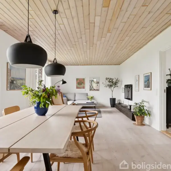 Spisebord med træstole står i en lys spisestue. Loftet er i lyst træ. Sorte lamper hænger over bordet. I baggrunden ses en sofa, planter og kunst på væggene.