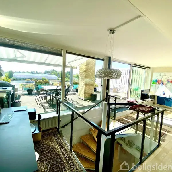 Et moderne stueområde med en trappe markeret af glasrækværk. Der er et skrivebord med en computer og udsigt til en terrasse med havemøbler og markise.