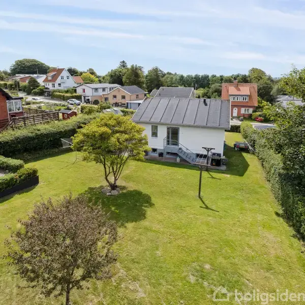 Et hvidt hus med sort tag står i en velholdt grøn have med træer og en hækkant. Flere andre huse ses i baggrunden i et fredeligt boligkvarter.