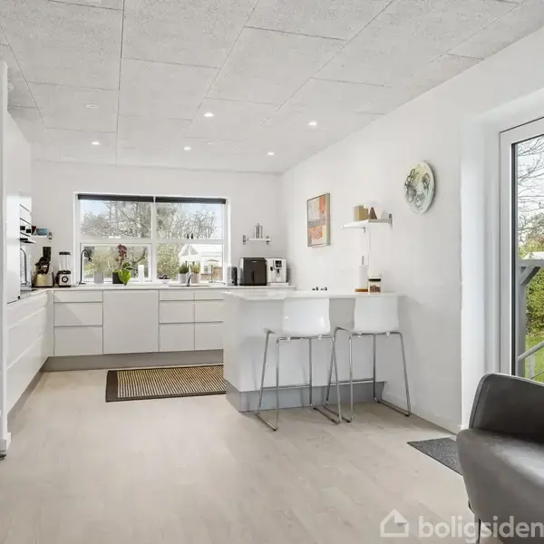 Køkken med hvide skabe og en stor ø, på baggrund af et stort vindue med udsigt til have. Indrettet med barstole og en lænestol.