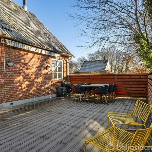 En terrasse med træbrædder har et sæt gule stole og et sort udendørs bord med stole. Den er omgivet af et lavt hegn ved et murstenshus med skygger på væggen.