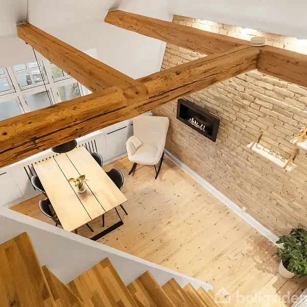 Et rum med trægulv og synlige loftsbjælker, indrettet med et spisebord, stole og en lænestol. Mod væggen er en elpejs indbygget i en murstensvæg. En plante står ved siden af.