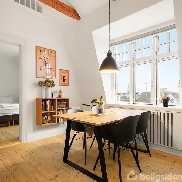 Et spisebord med sorte stole står under en hængende lampe i en lys stue. Et stort vindue slipper dagslys ind; væggen har kunst og en boghylde.