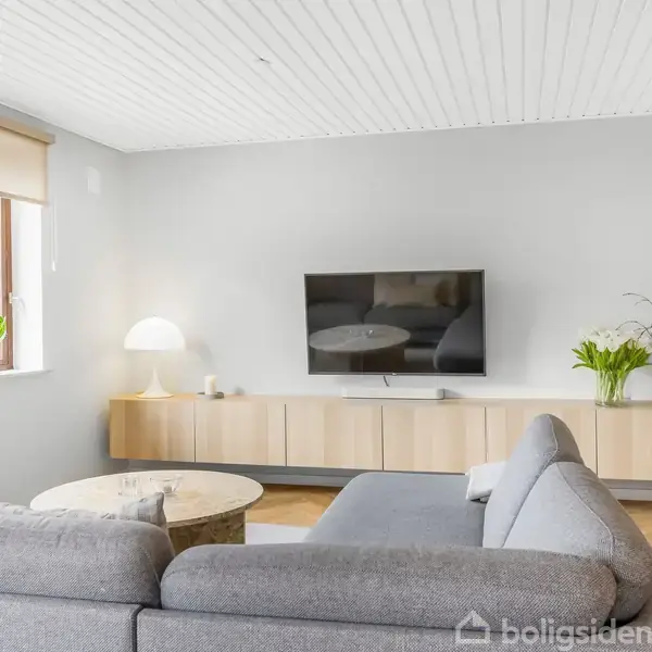 Gråt sofaarrangement står foran en vægmonteret fladskærm i en lys stue. En lav reol med vaser og en lampe står ved tv'et. Vindue til venstre med udsyn til grønne planter.