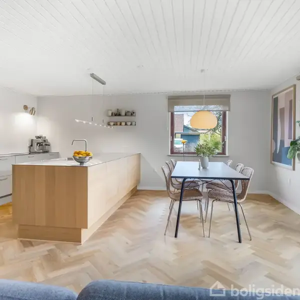 Køkken med en træø med frugtskål. Til venstre er der moderne husholdningsapparater. Spisebord med stole står ved vinduet. Trægulv og hvidt loft skaber en lys atmosfære.