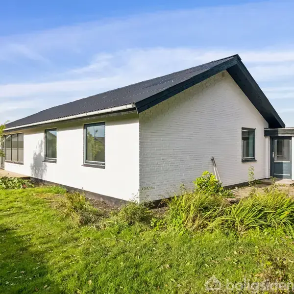 En hvid murstenshus med sort tag står i en have med grøn græsplæne og buske. Vinduerne er kvadratiske, og huset har en lille flisebelagt indgangsvej foran.