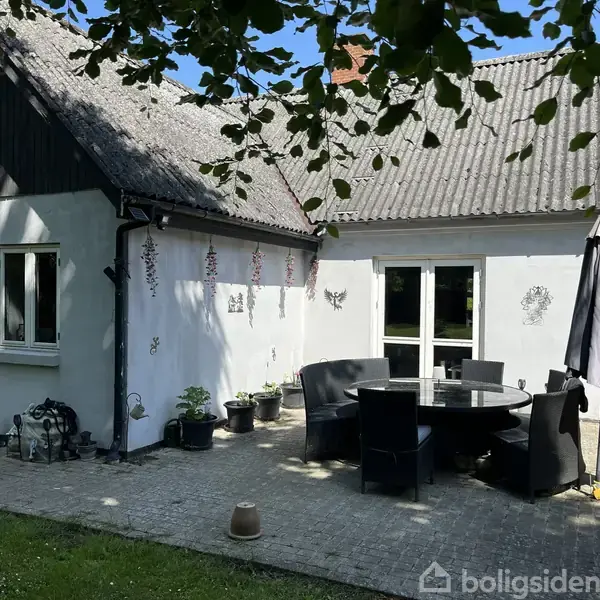 Huset har en hvid facade med skråt tag. Foran ses en terrasse med et sort rundt bord og stole. Haveparasollen står ved siden af. Omkring er der græs og dekorationer på væggen.