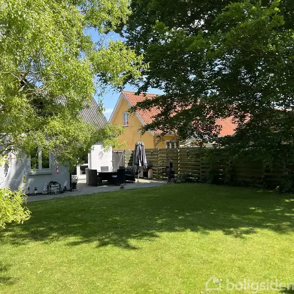 Hus med terrasse møbleret med sofagruppe og parasol, omgivet af grønne træer og en velplejet græsplæne. I baggrunden ses to farverige huse.